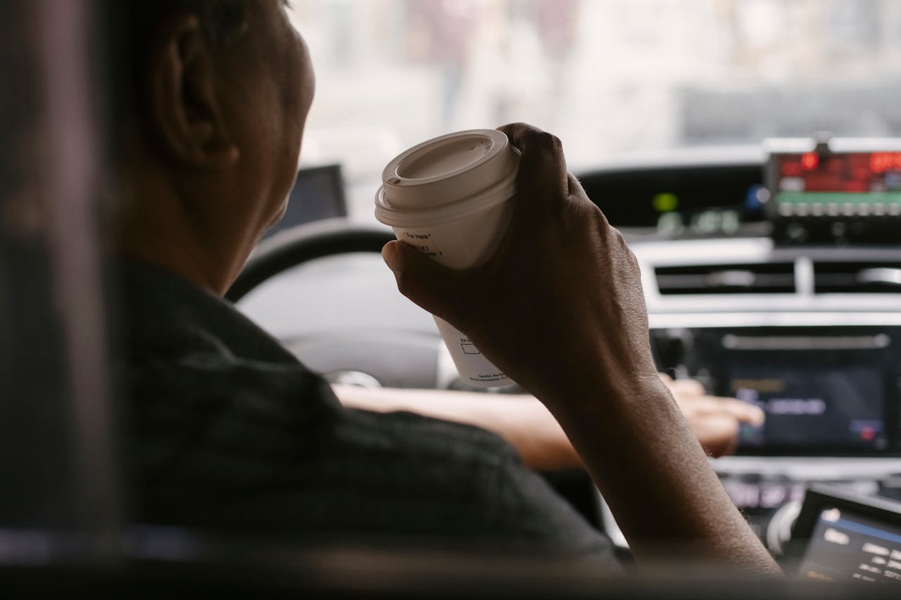 services-01 Back view of unrecognizable male drinking coffee in drivers seat at wheel while looking away on urban background in soft daylight