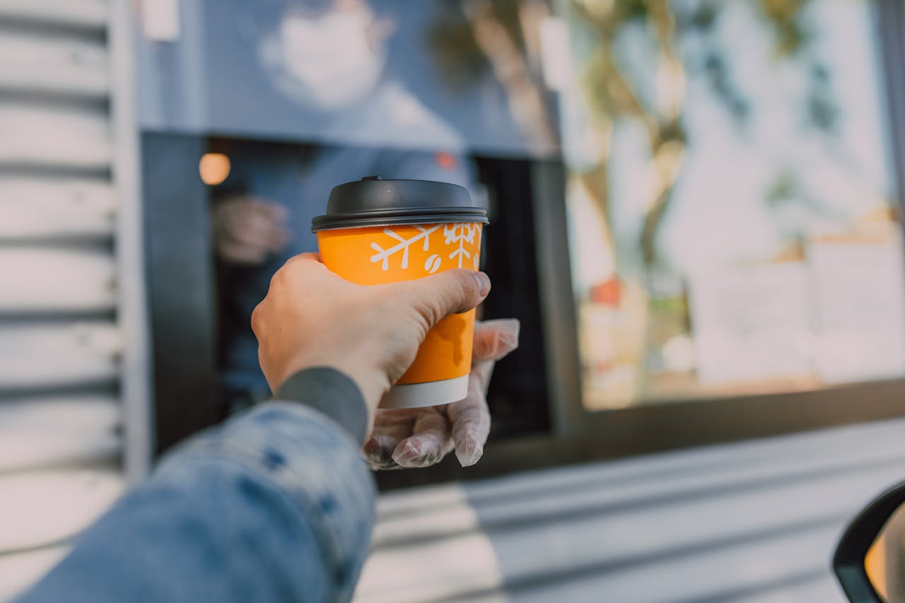 hero-img-02 Hand reaching out for a hot coffee cup at a drive-thru window, depicting everyday convenience.